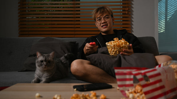 Happy young Asian man watching TV and eating popcorn while sitting with a lovely cat on a cozy sofa.